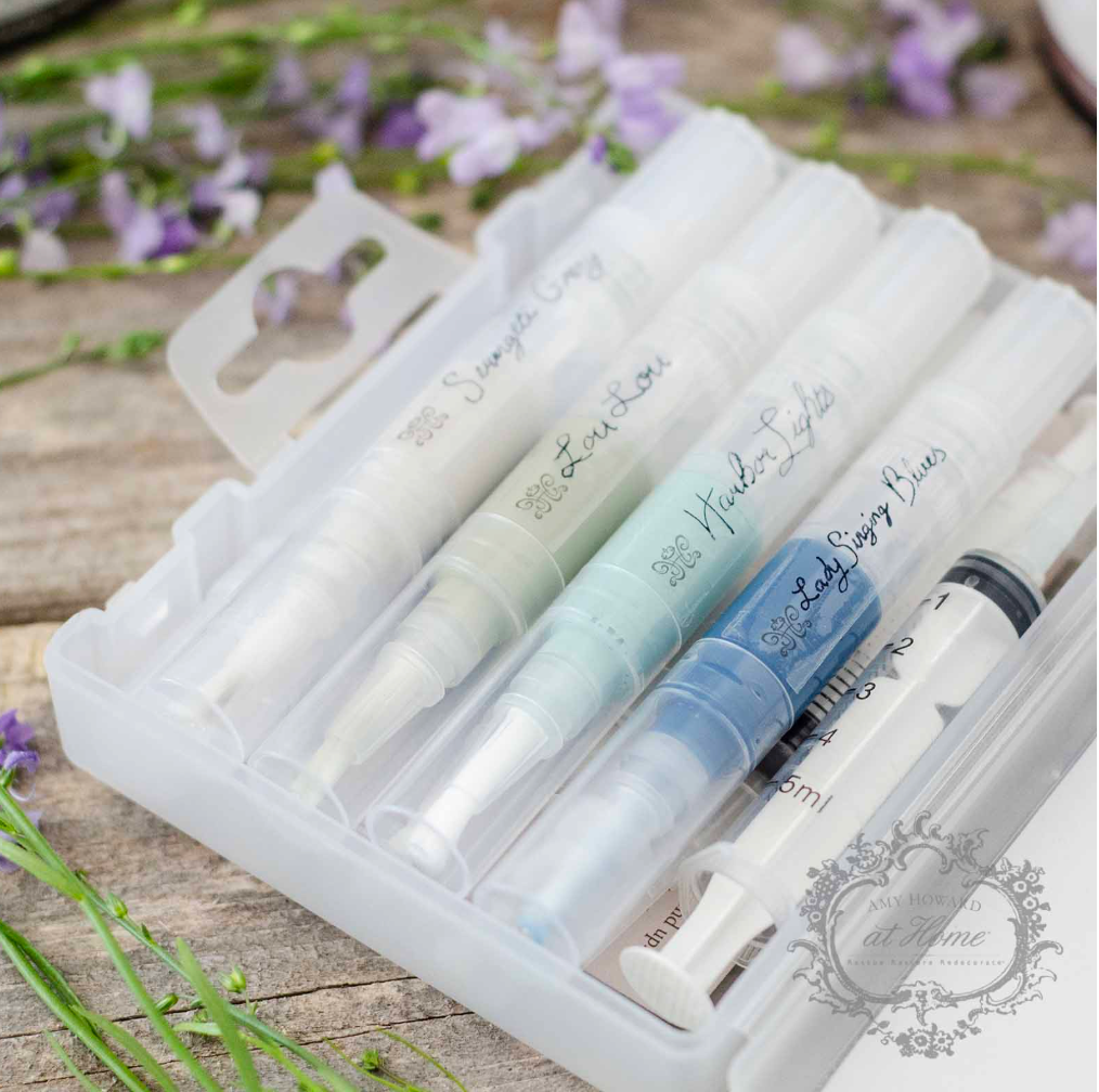 Set of paint tubes in a clear case on a wooden surface with flowers in the background